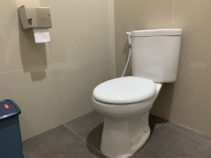 Un inodoro estándar con tanque y asiento blancos, un dispensador de toallas de papel y un cesto de basura.