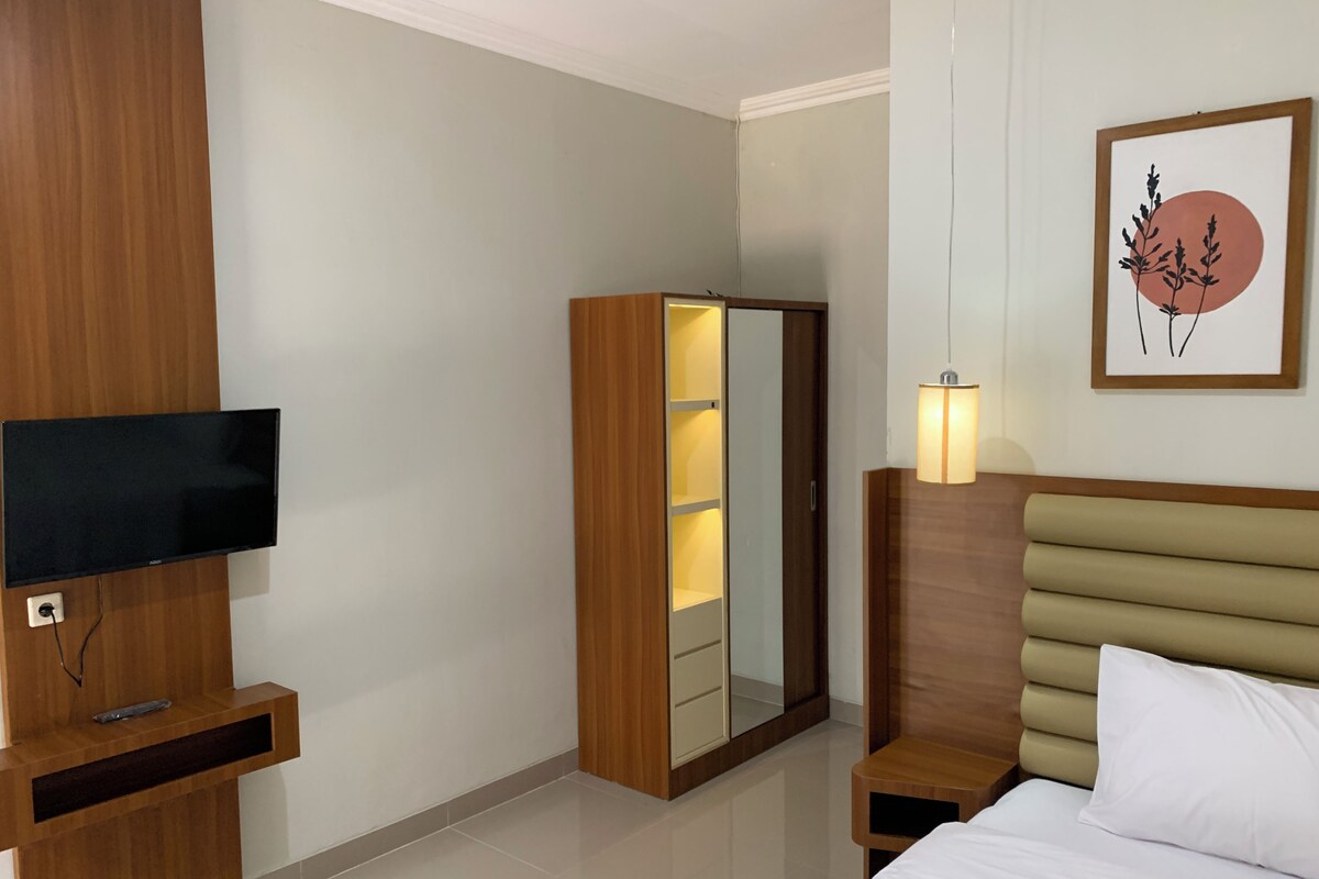 A hotel room with a bed, a TV mounted on a wooden panel, a wardrobe, a bedside table with a lamp, and a framed artwork on the wall.