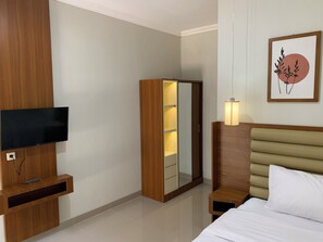 A hotel room with a bed, a TV mounted on a wooden panel, a wardrobe, a bedside table with a lamp, and a framed artwork on the wall.
