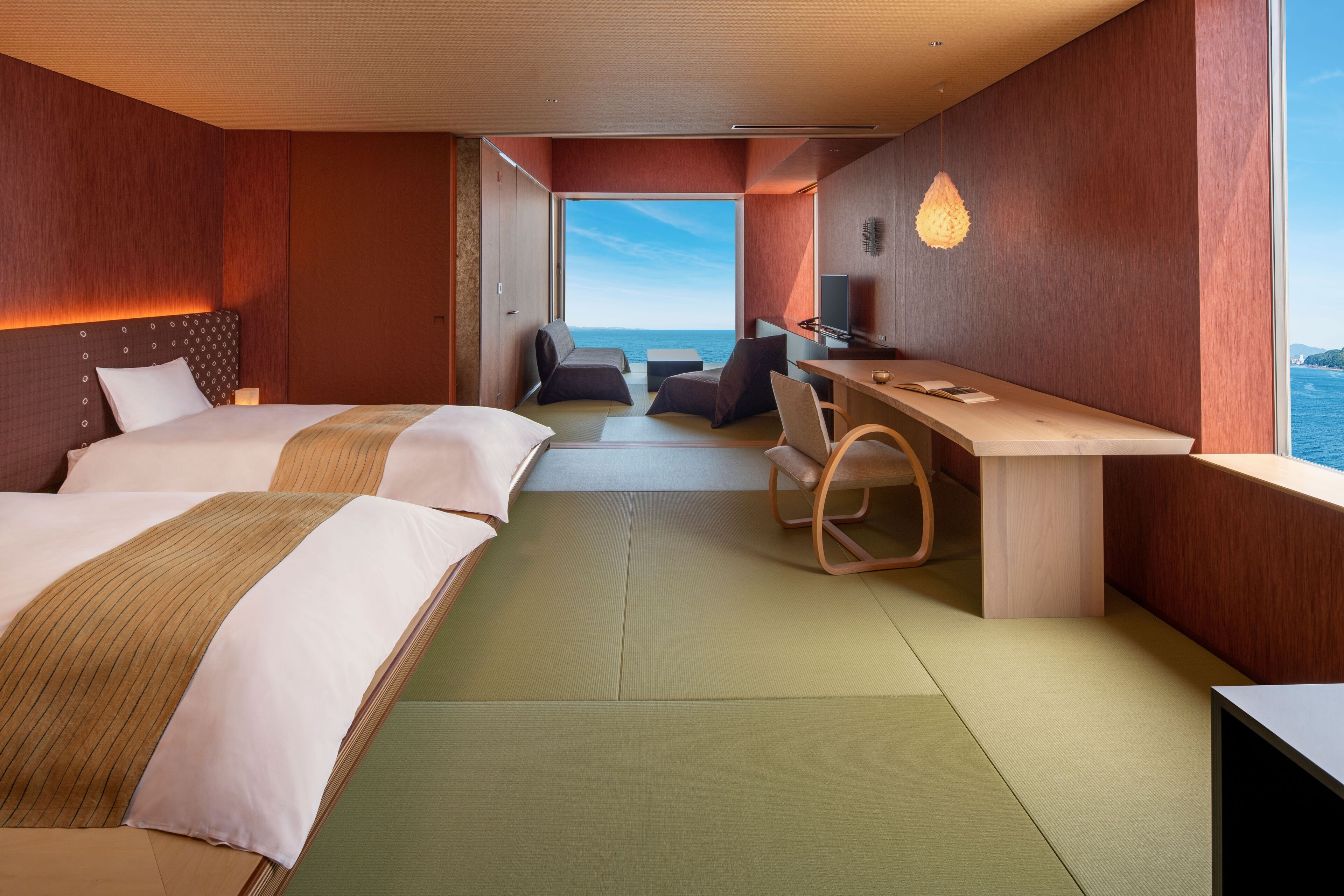 A modern hotel room with two beds, a desk, and a view of the sea.