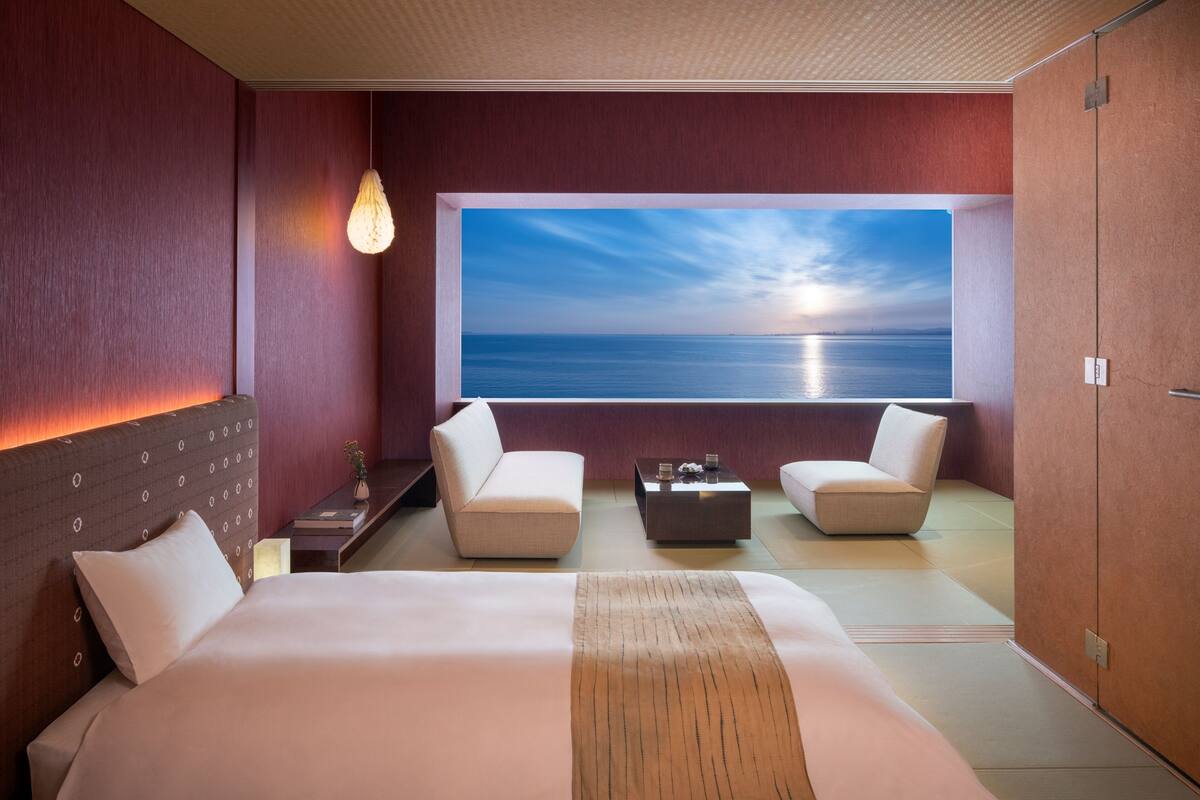 Een moderne hotelkamer met een groot bed, twee fauteuils, een salontafel en uitzicht op zee.