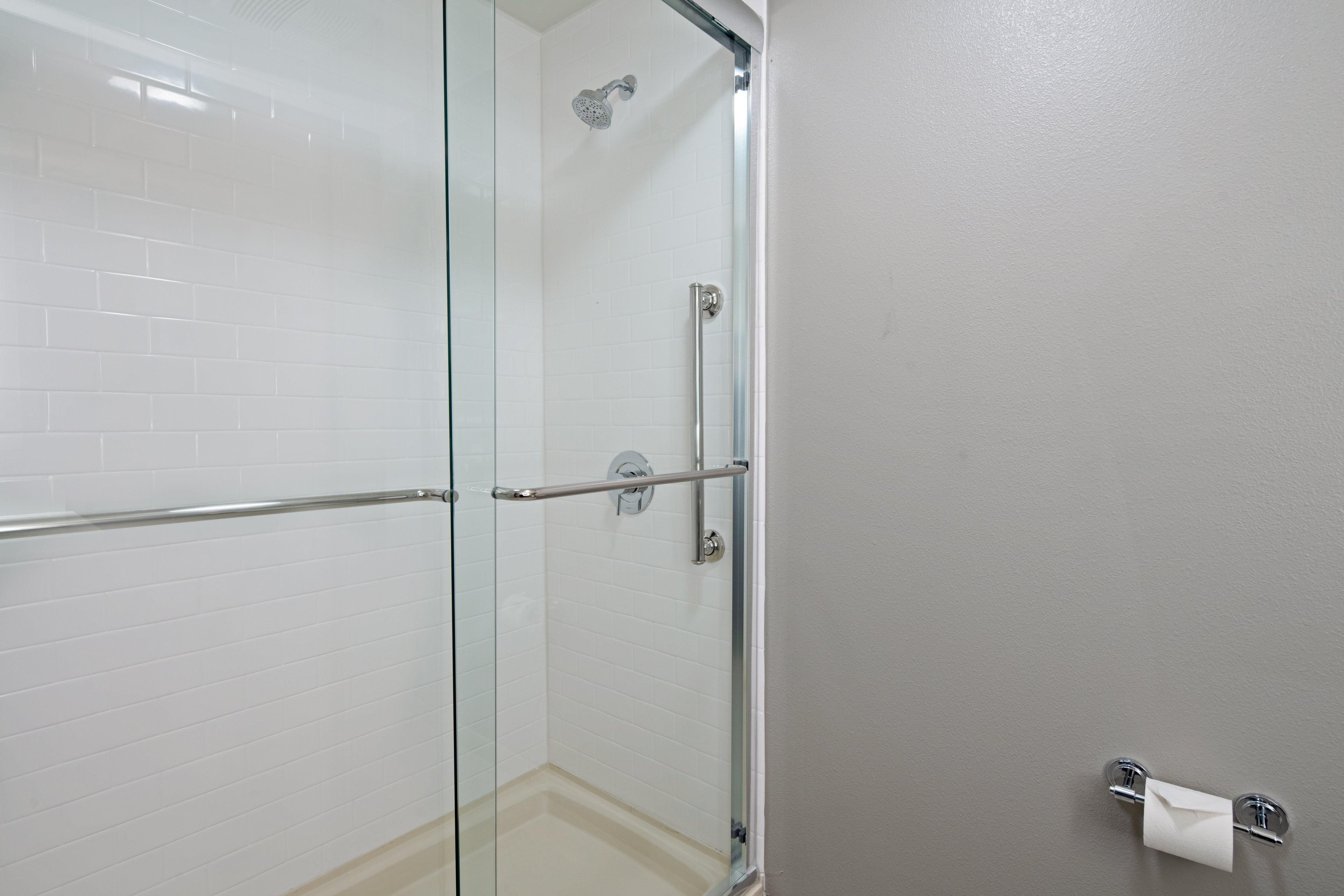 A glass-enclosed shower with a metal handrail, a white tiled wall, and a towel dispenser on the right.