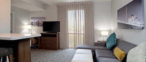 A modern hotel room with a flat-screen TV, a sofa, a small table, a desk, and a large window with curtains.