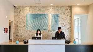 A reception desk with two staff members, a tiled wall with a geometric artwork, and a window to the right. - Staybridge Suites Long Beach Airport (Long Beach)