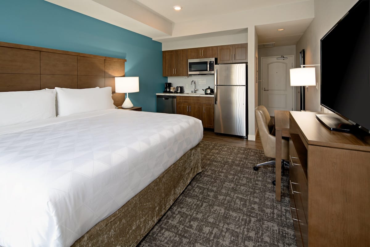 Quarto de hotel com cama grande, escrivaninha, cozinha compacta e televisão.