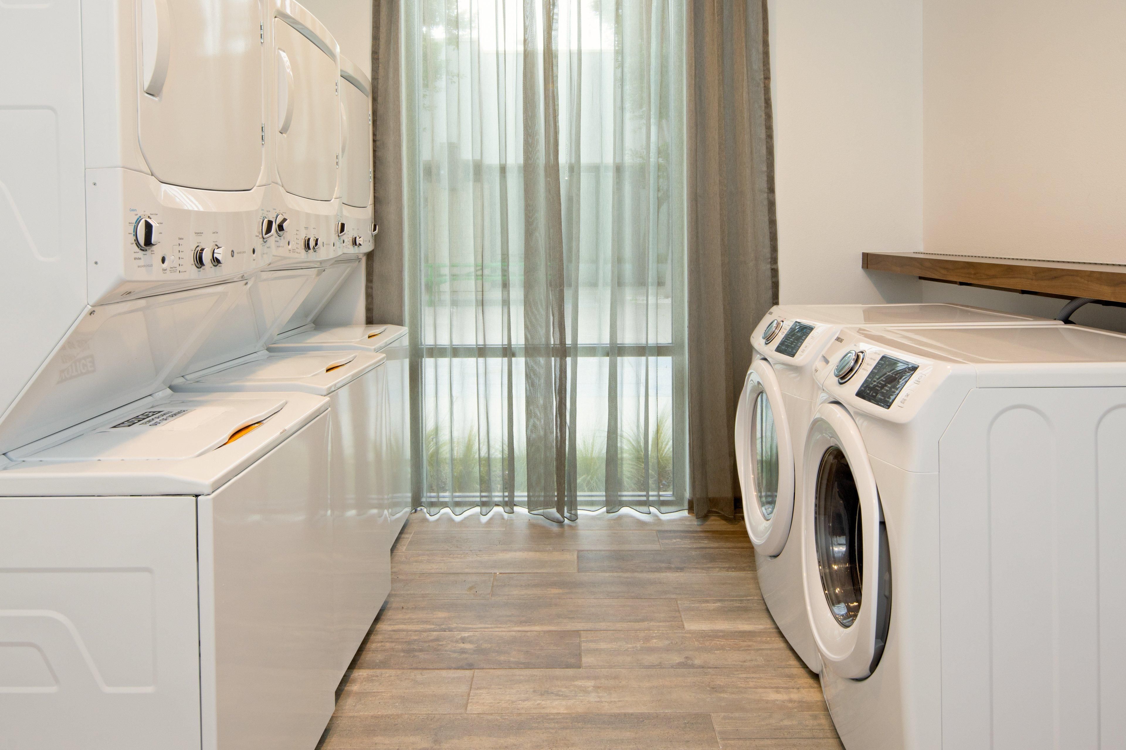 A laundry room with multiple washing machines and dryers, wooden flooring, and a large window with sheer curtains.