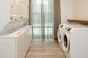 Een wasserette met meerdere wasmachines en drogers, houten vloeren en een groot raam met dunne gordijnen.
