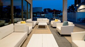 Ein moderner Außenbereich mit weißen Sofas und Tischen, großen Fenstern und Blick auf die Stadt bei Dämmerung.
