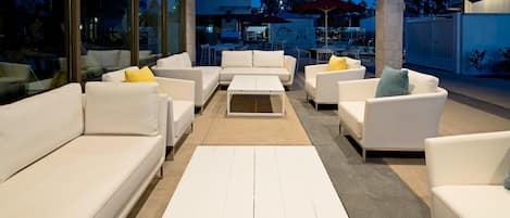 A modern outdoor seating area with white sofas and tables, large windows, and a view of the cityscape at dusk.