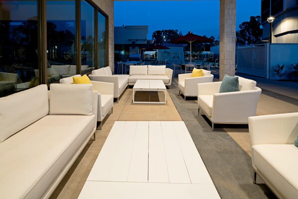 Un espace extérieur moderne, aménagé avec des canapés et des tables blancs, de grandes fenêtres et une vue sur le paysage urbain au crépuscule.