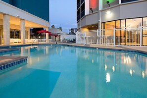 Une piscine entourée d’une clôture blanche, avec des chaises longues et un bâtiment doté de grandes fenêtres.