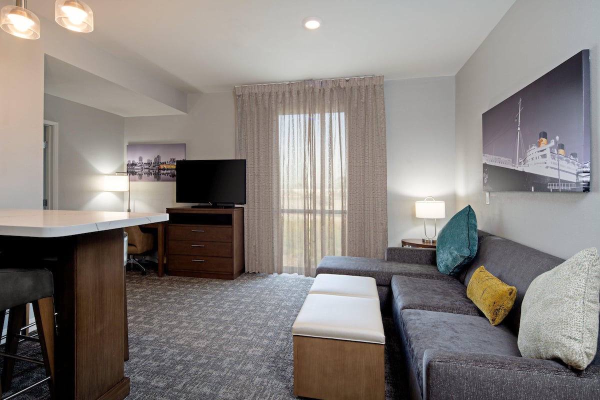 Quarto de hotel moderno com televisão de tela plana, sofá, mesa pequena, escrivaninha e uma grande janela com cortinas.