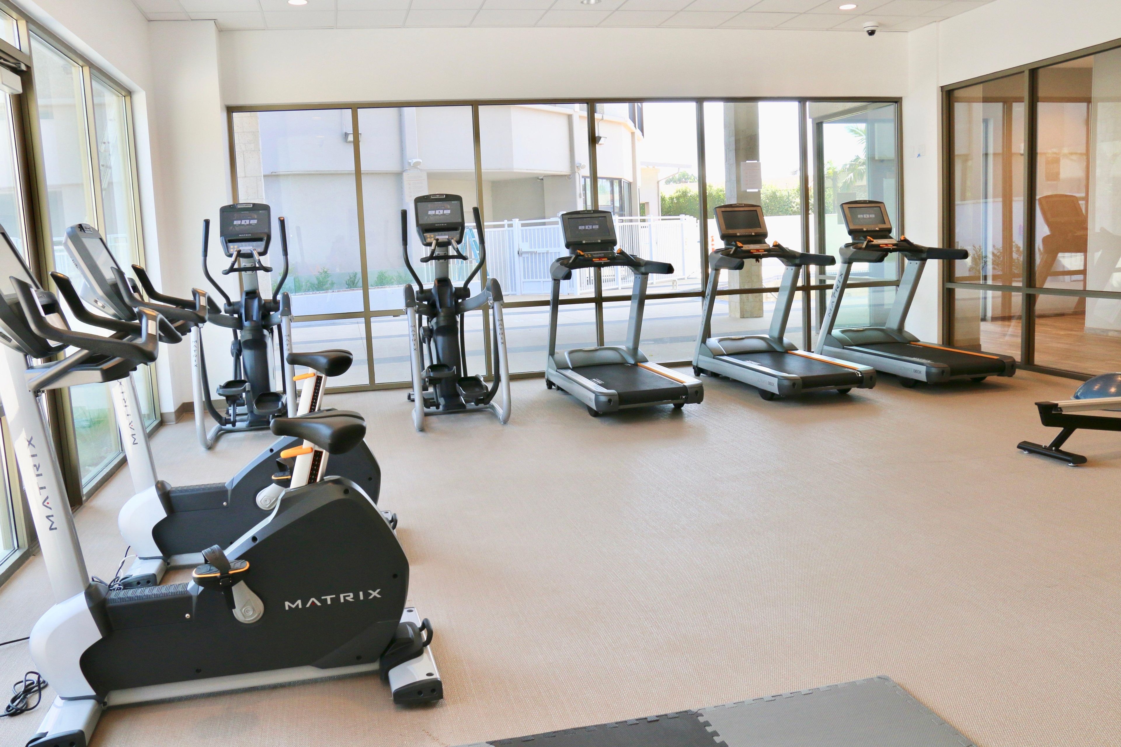 A gym with various exercise machines, large windows, and a view of the outside.