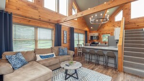 A cozy cabin interior with a wooden interior, a sofa, a coffee table, a kitchen area, and a staircase. - Family Lake Retreat w/ Fire Pit & Hot Tub (Swanton)