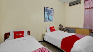 Ein Hotelzimmer mit zwei Betten, jedes mit einer roten und weißen Decke, und einem roten Kopfkissen mit dem Logo "OYO". Es gibt einen Holztisch, einen Stuhl und eine Wand-Klimaanlage. An der Wand hängt ein gerahmtes Bild von einem Baum.