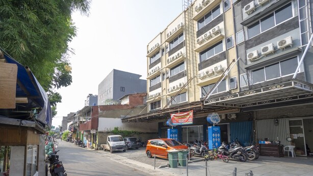 A street with parked vehicles, including motorcycles and cars, and a building with multiple air conditioning units on the facade. - OYO 3754 Prosperity Mansion (Jakarta)
