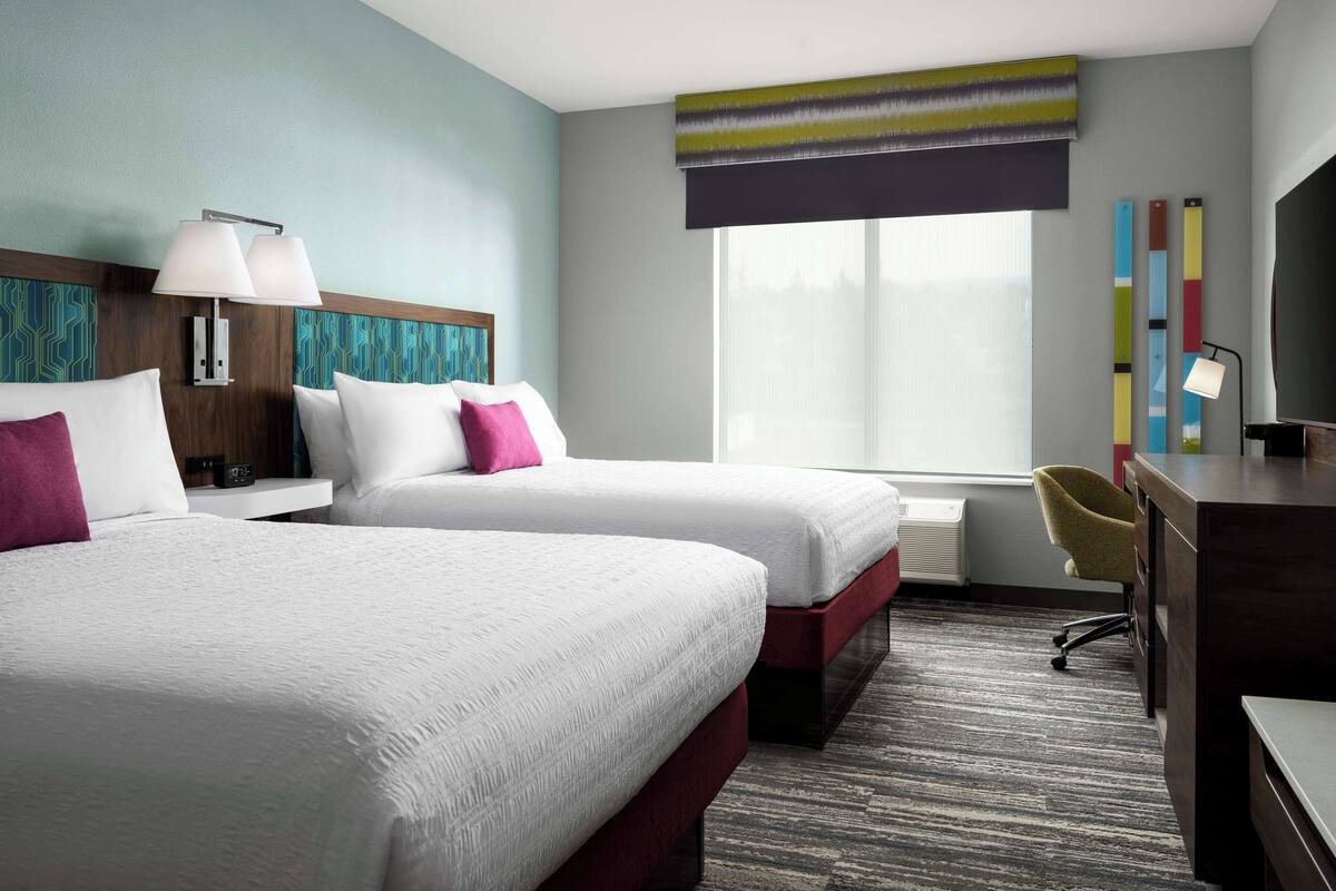 A hotel room with two beds, a desk, a chair, a TV, and a window with blinds.