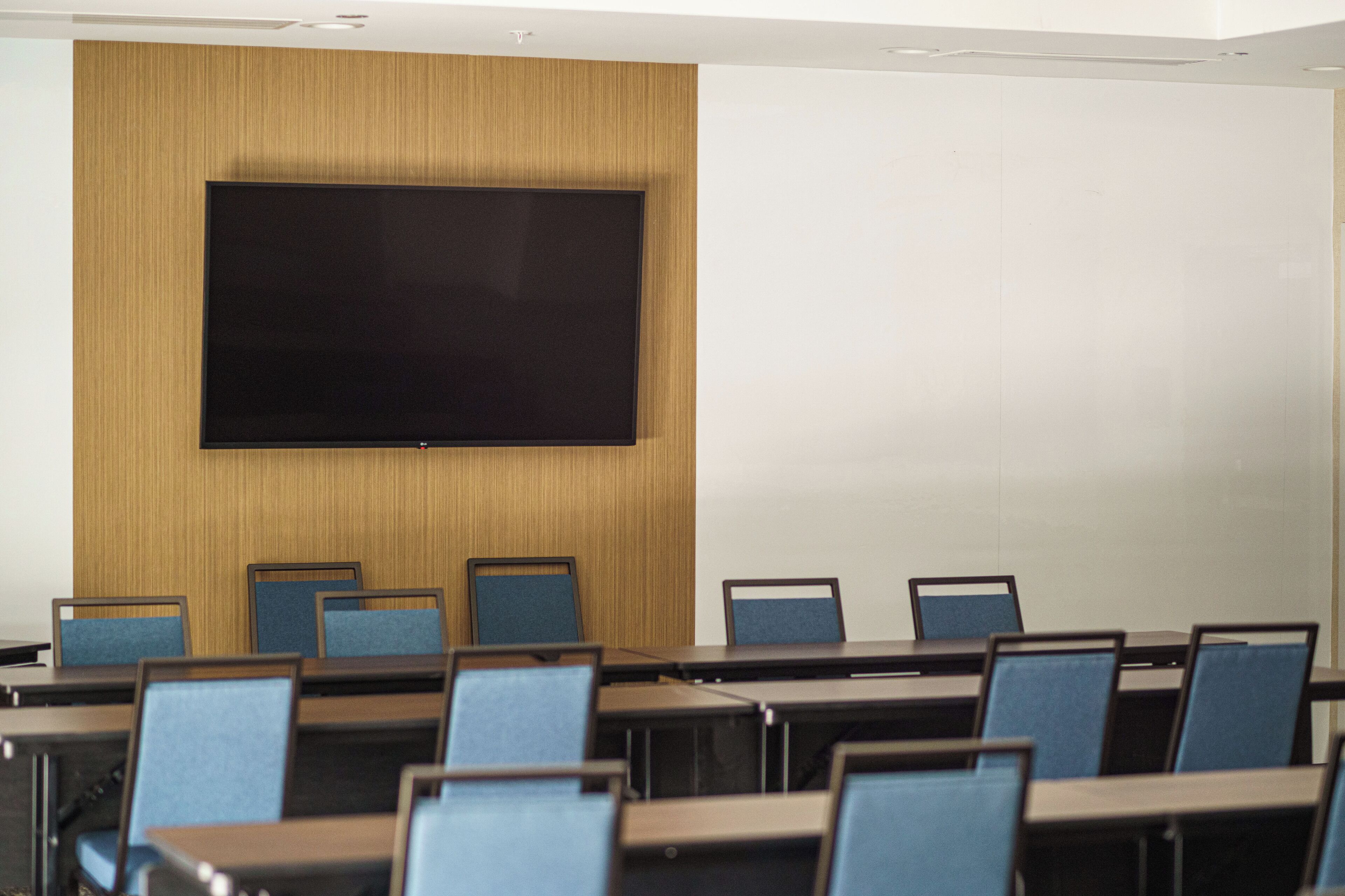 A conference room with a large flat-screen TV mounted on a wooden wall, rows of blue chairs, and tables.