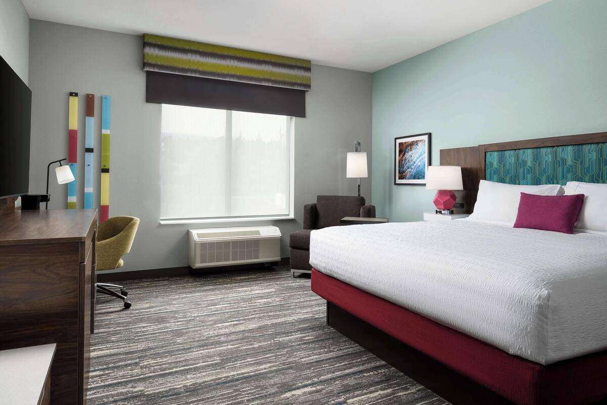 A hotel room with a large bed, a desk with a chair, a TV, a window with blinds, and a painting on the wall.