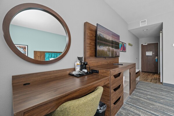 A hotel room with a wooden desk, a large mirror, a flat-screen TV, and a painting on the wall.