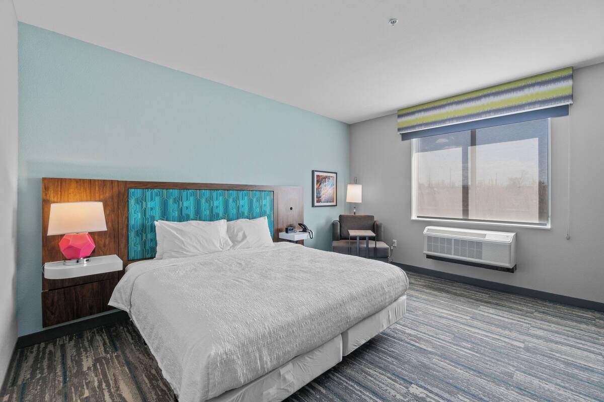 A hotel room with a large bed, a nightstand with a lamp, a chair, a window with blinds, and an air conditioning unit.