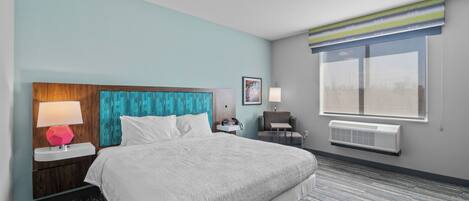 A hotel room with a large bed, a nightstand with a lamp, a chair, a window with blinds, and an air conditioning unit.
