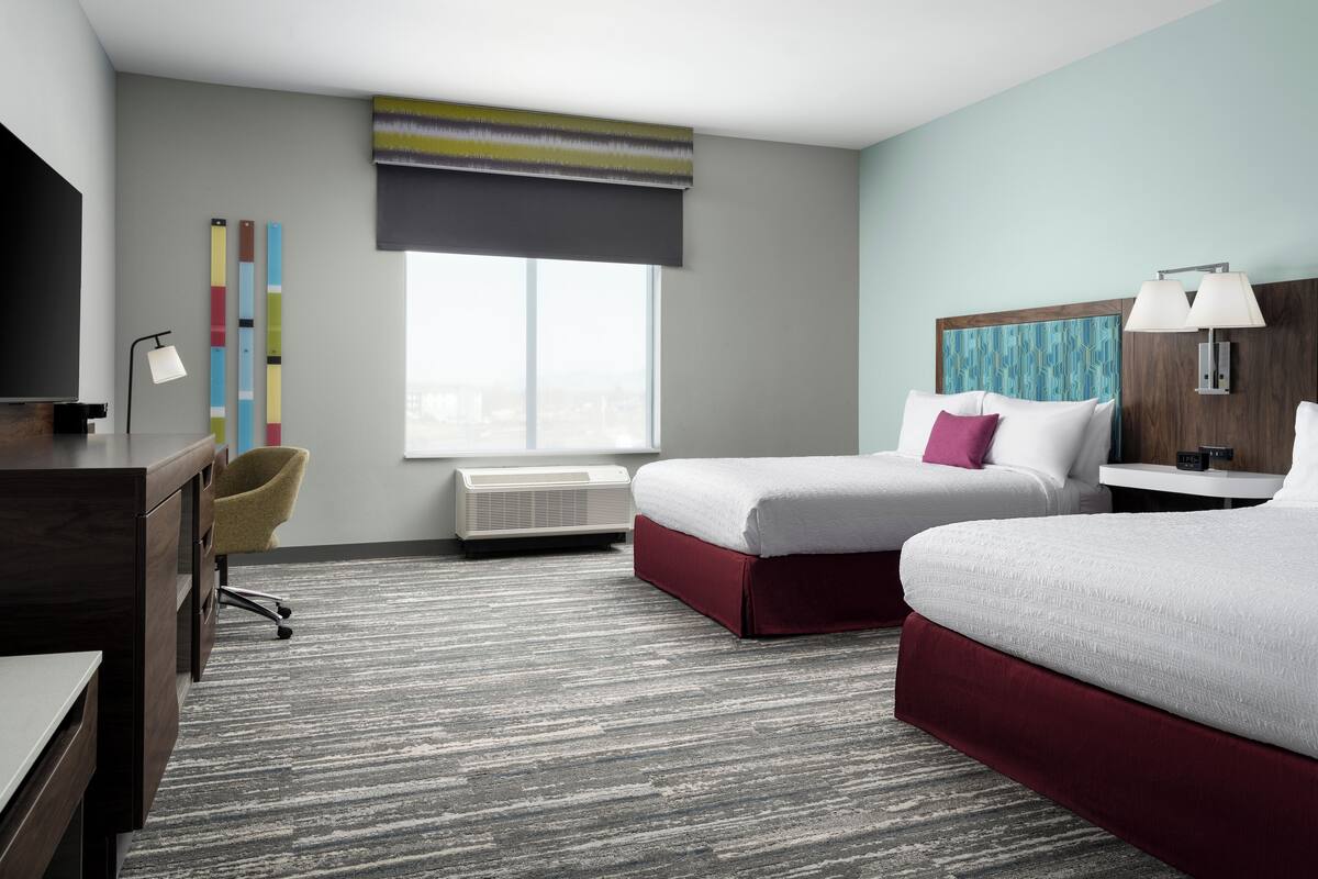 A hotel room with two beds, a desk, a chair, a TV, a lamp, and a window with a view.