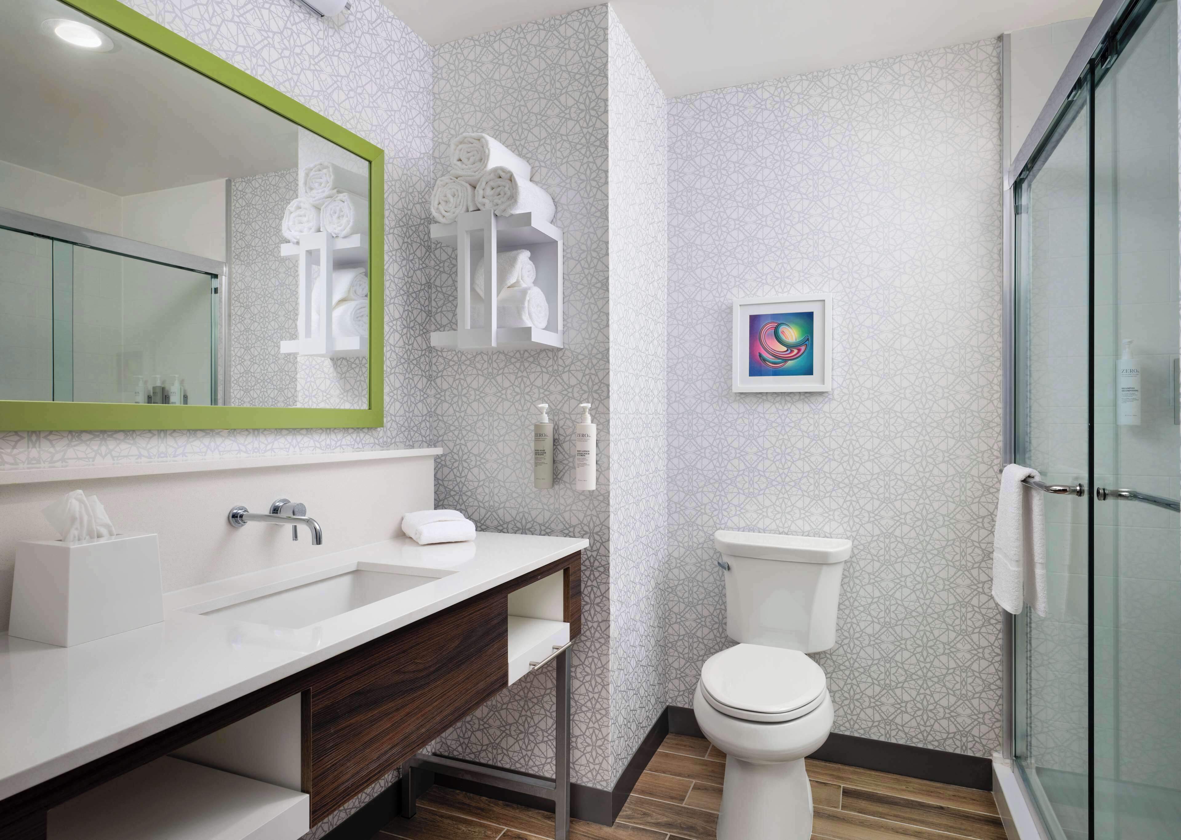 Un baño moderno con un lavamanos blanco, un espejo y un inodoro. Hay una zona de ducha con puerta de vidrio, y las paredes presentan un diseño geométrico.