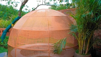 Una estructura circular tejida con techo de malla, rodeada de follaje y un muro de ladrillo.