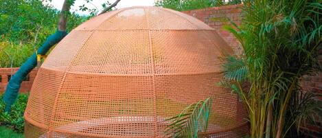 A round, woven structure with a mesh roof, surrounded by greenery and a brick wall.