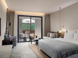 A hotel room with a bed, a desk, a chair, a table, and a view of greenery outside.