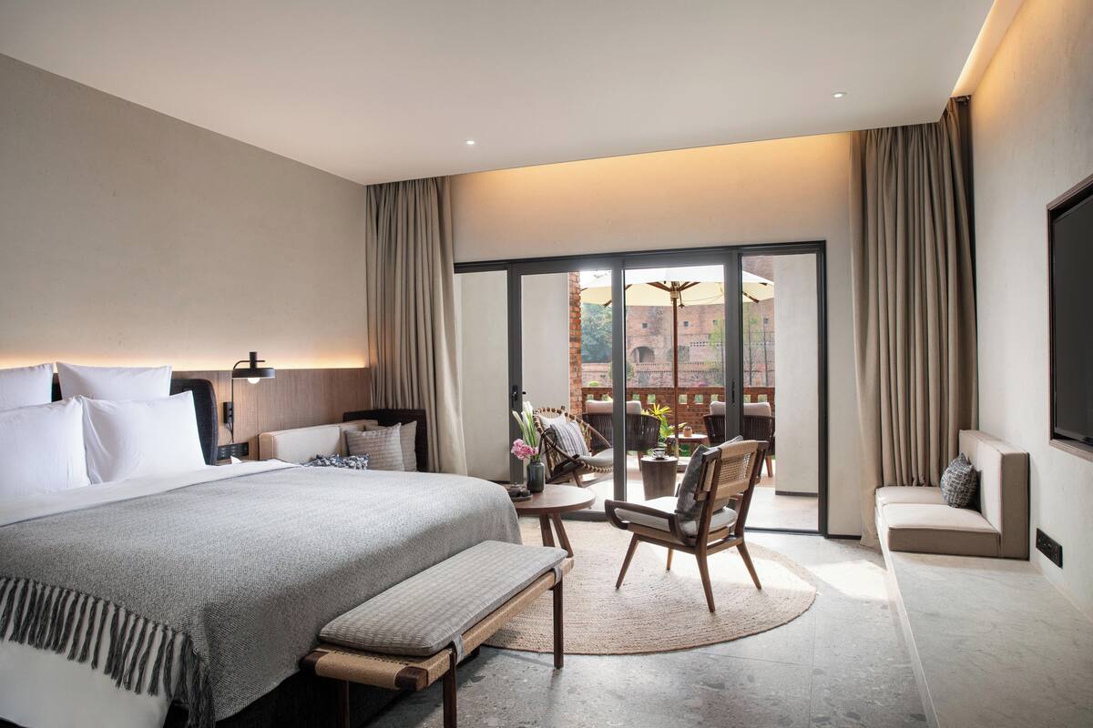 A modern hotel room with a large bed, a sofa, a dining table, and a view of the outside.