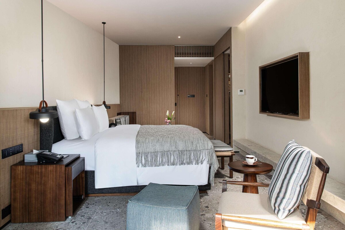Habitació d'hotel moderna amb llit, taulell de nit, làmpada, cadira i taula petita amb una tassa.