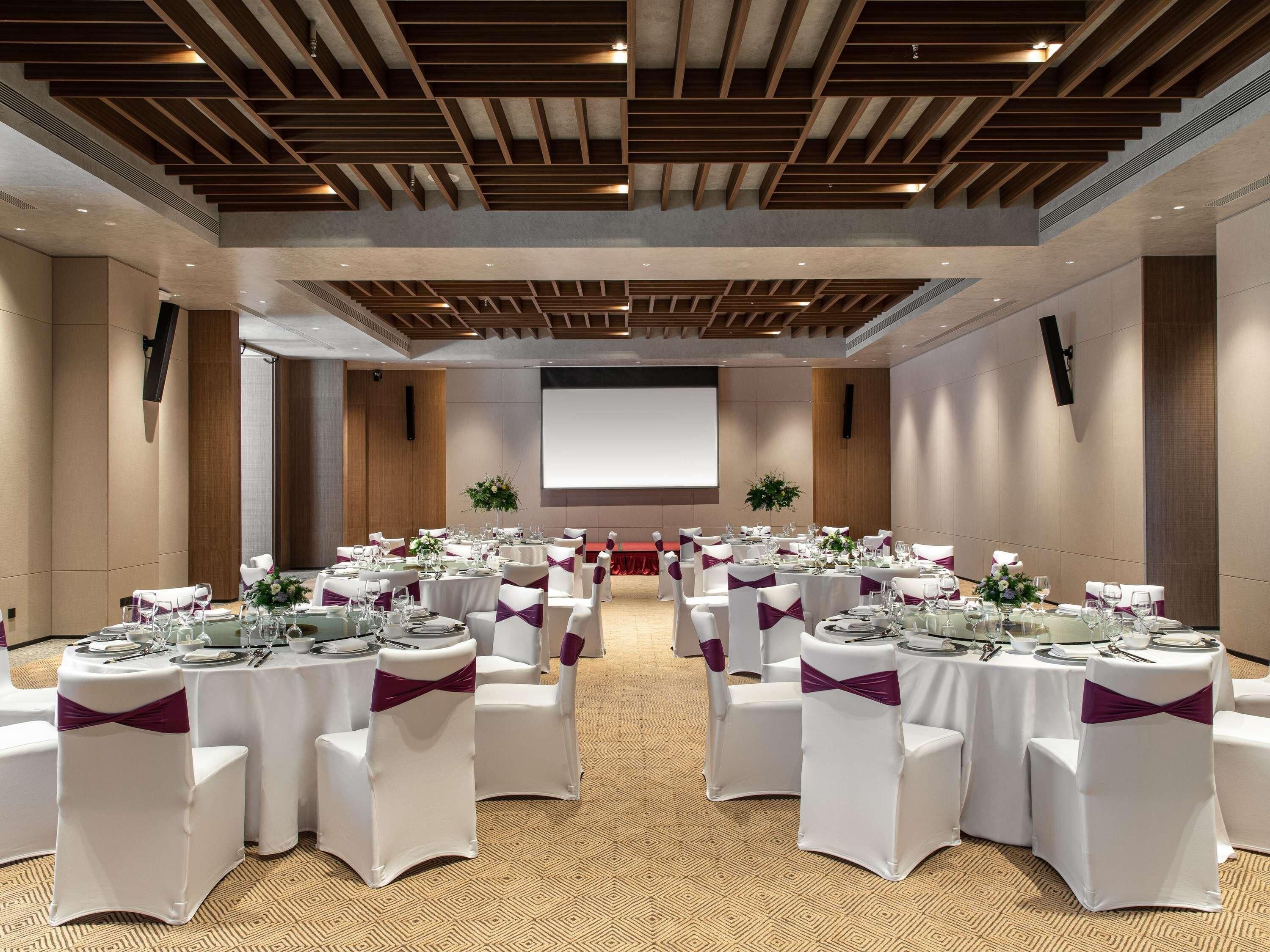 Een vergaderzaal met witte tafels, stoelen en een groot scherm.