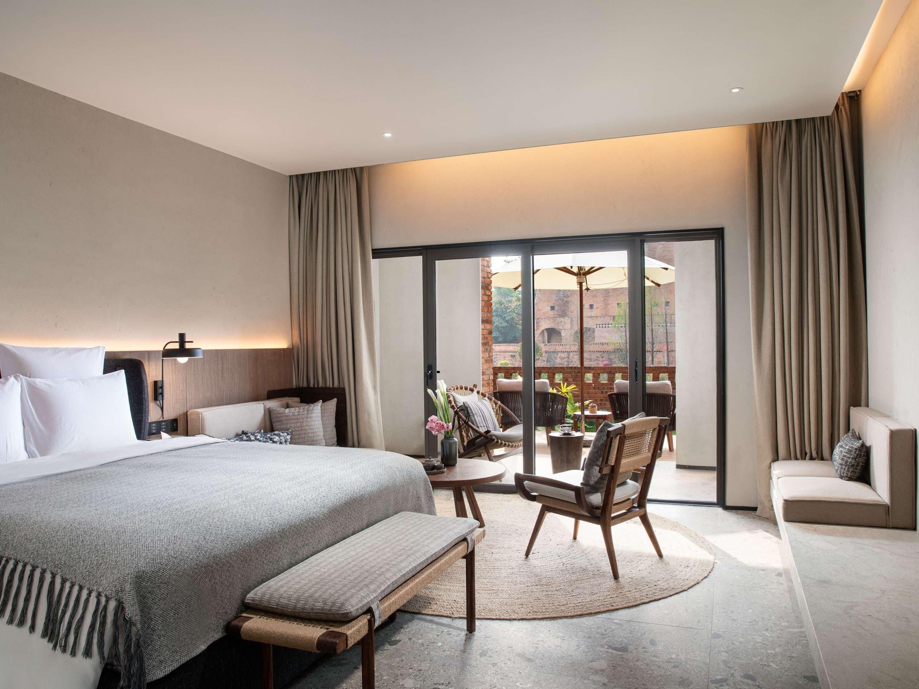 Una habitación de hotel moderna con una cama grande, una zona de estar con sillas, una mesita y vistas a un patio exterior a través de un ventanal.