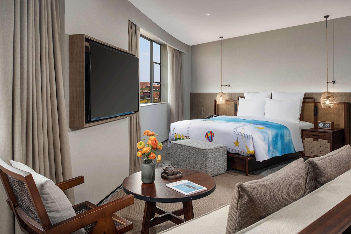 Habitación de hotel con cama, televisor, una mesa con un florero, un sofá y una silla.