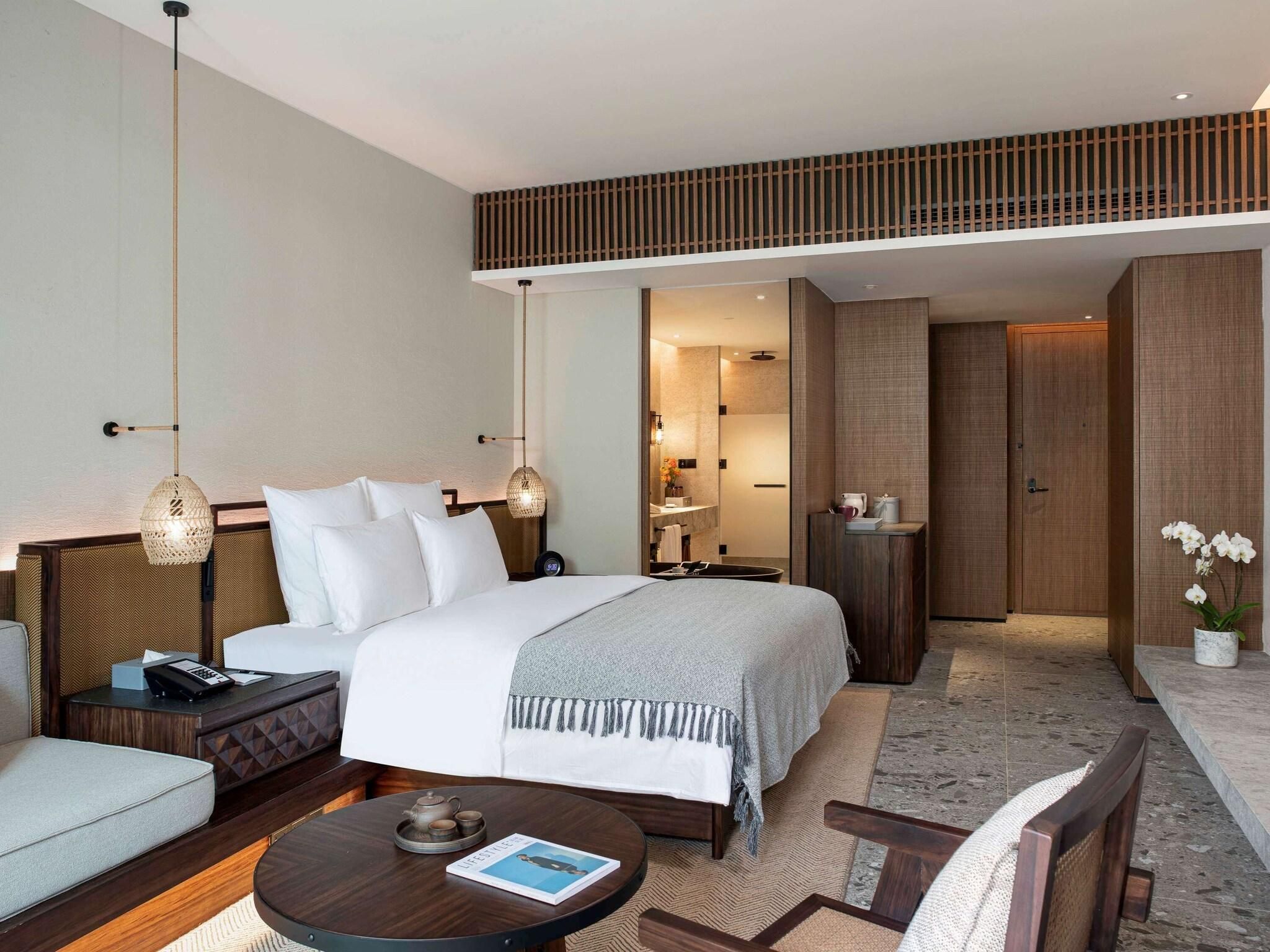 A modern hotel room with a large bed, a sitting area with a coffee table, a small table with a teapot, and a mirror reflecting the bathroom.