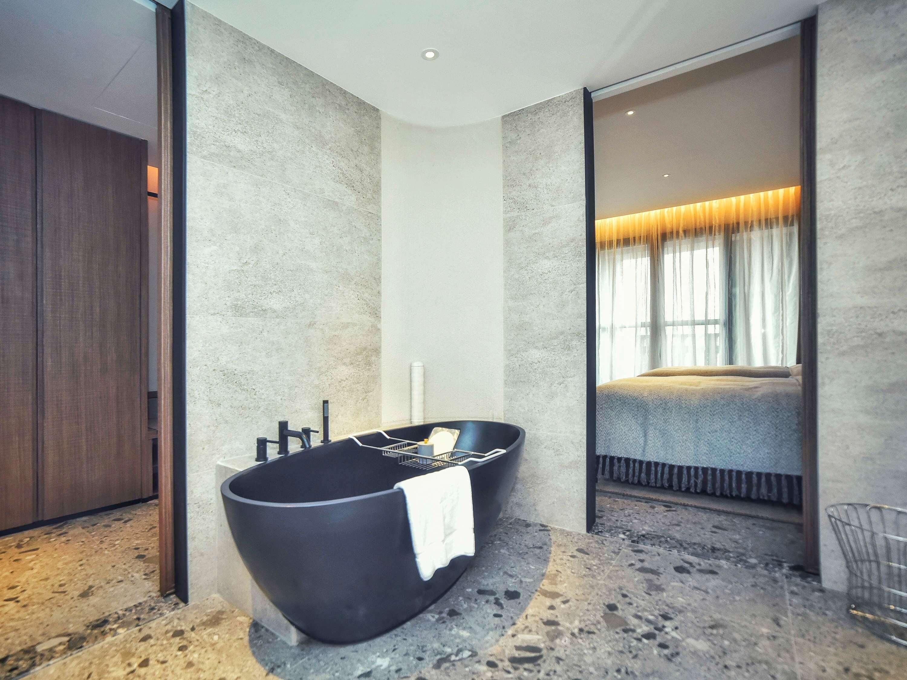 Un baño moderno con una bañera negra, un espejo grande y vistas hacia un dormitorio.