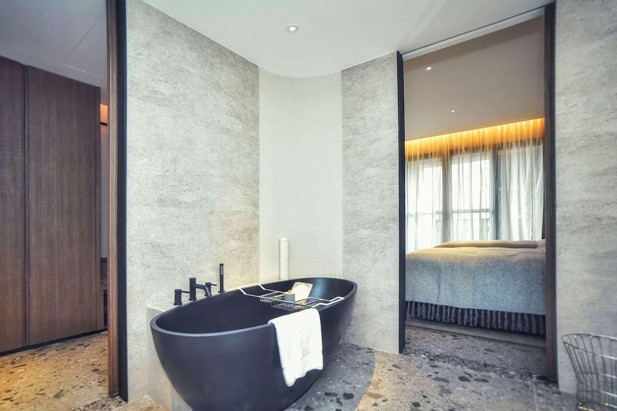 A modern bathroom with a black bathtub, a large mirror, and a view into a bedroom.