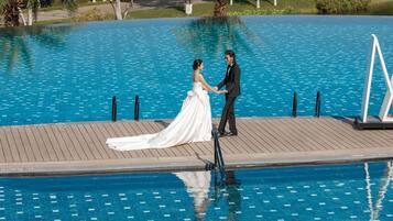 A couple holding hands on a wooden dock by a clear blue pool with a view of trees and a road in the background.
