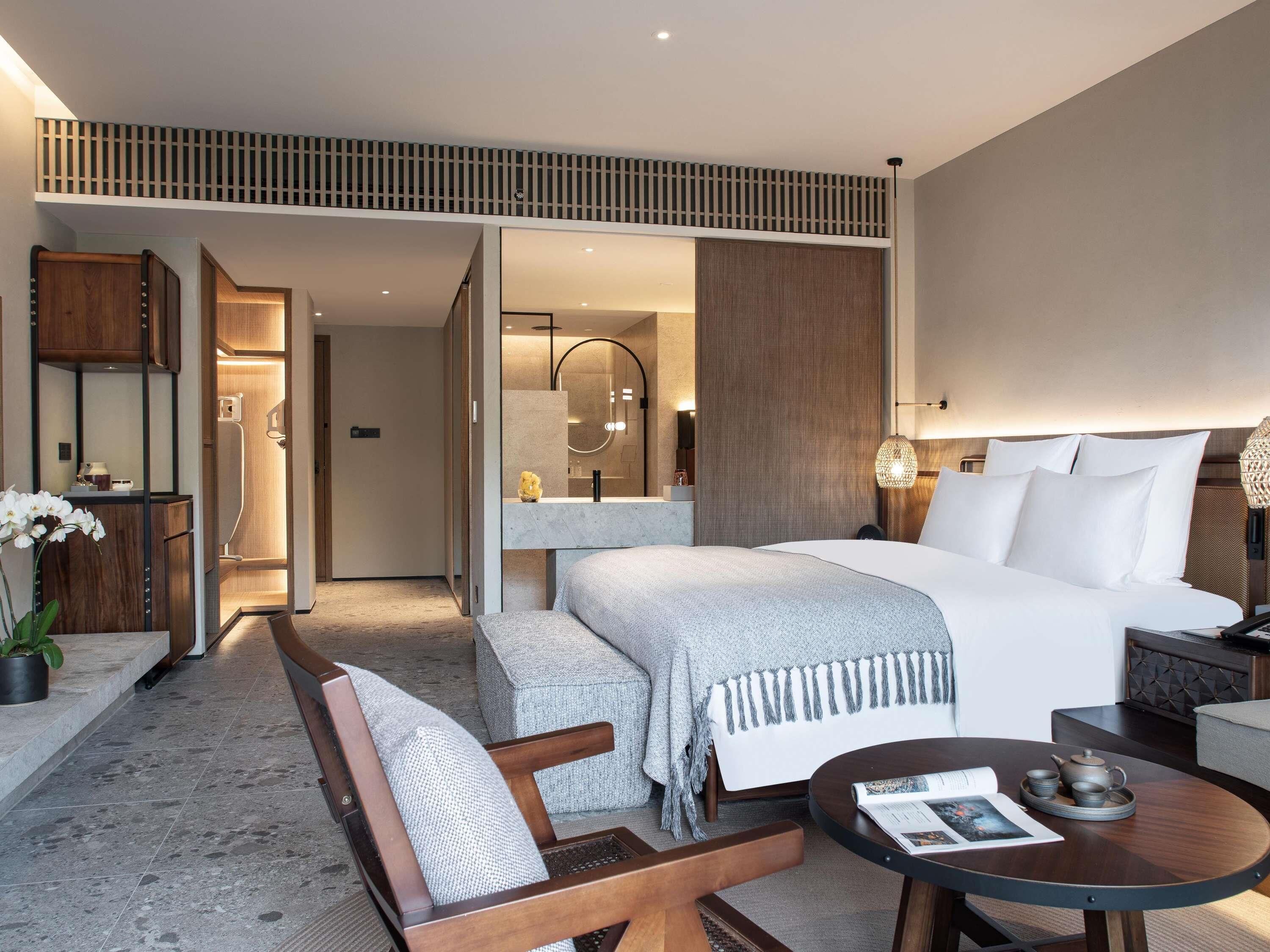 Una habitación de hotel moderna con una cama grande, una zona de estar con un banco y una silla, una mesita redonda con una revista y un baño con ducha y espejo.