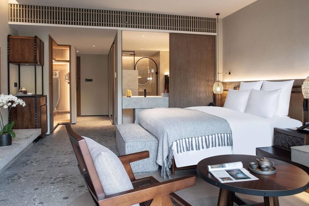 A modern hotel room with a large bed, a sitting area with a bench and chair, a small round table with a magazine, and a bathroom with a shower and mirror.