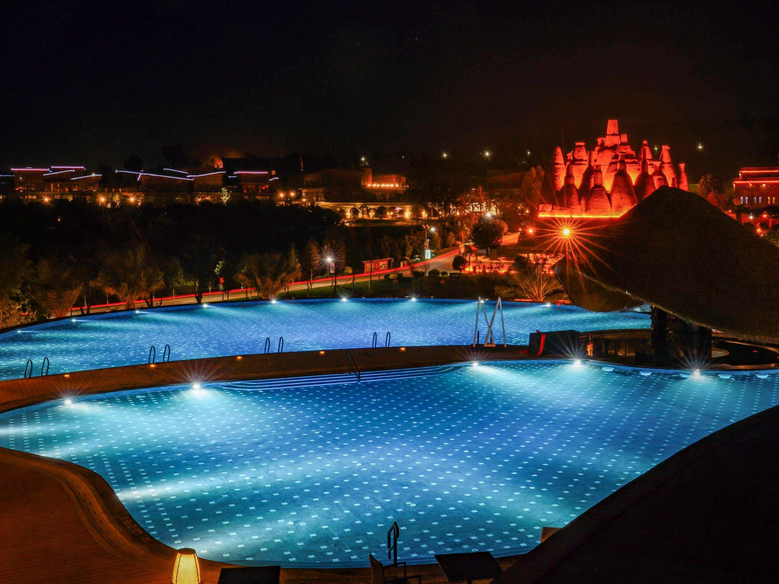 Vista nocturna de una piscina rodeada de edificios y una gran estructura iluminada al fondo.