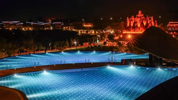 Vista nocturna de una piscina rodeada de edificios y una gran estructura iluminada al fondo.