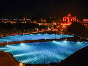 Vista nocturna de una piscina rodeada de edificios y una gran estructura iluminada al fondo.