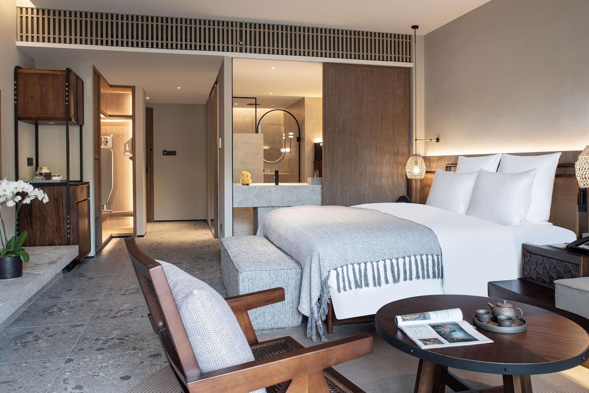 Una habitación de hotel moderna con una cama grande, una zona de estar con una silla y una mesa, un baño con ducha y espejo, y un diseño de techo de madera.