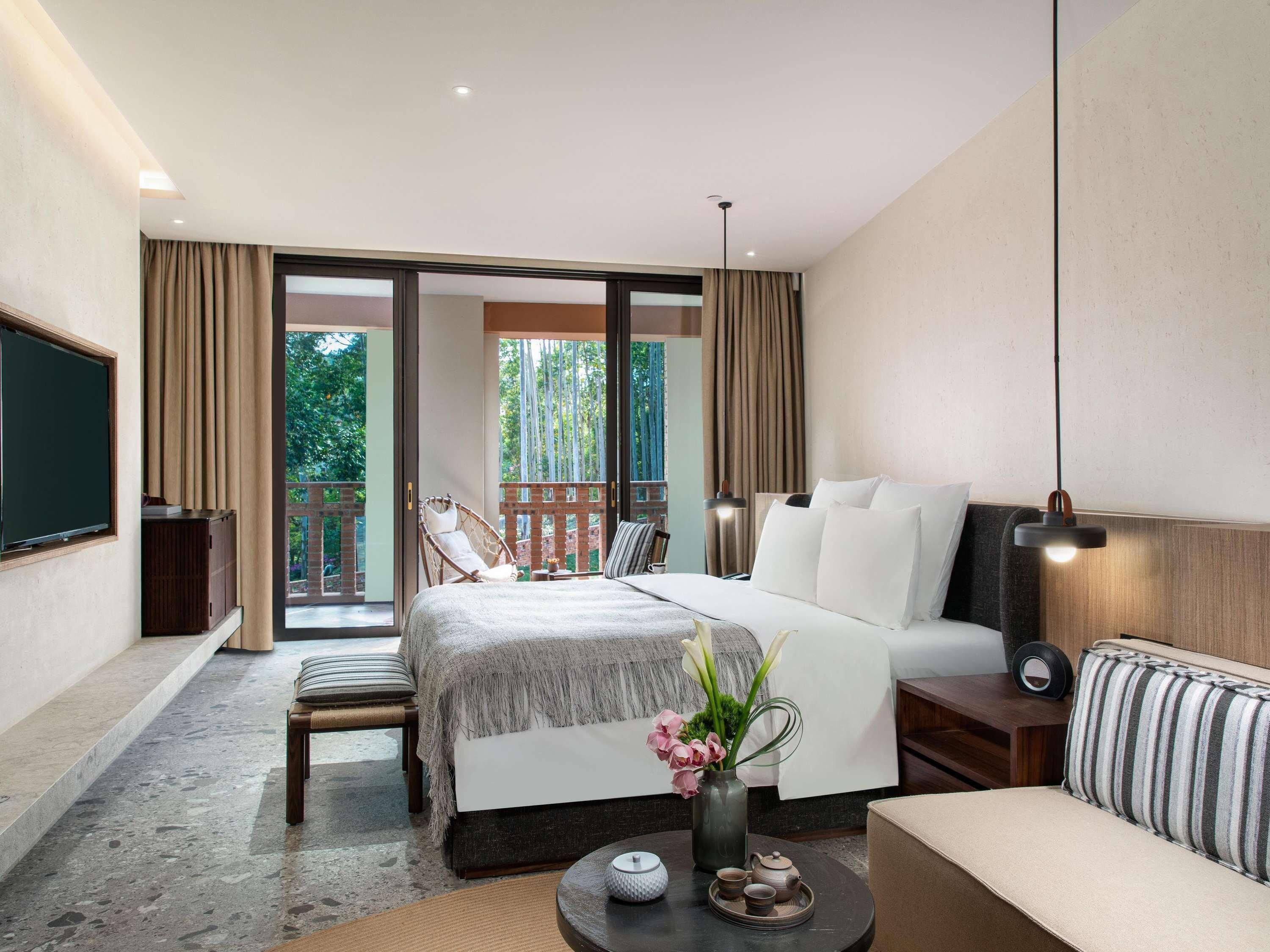 Habitación de hotel con cama, sofá, televisor, balcón y mesa con un florero y teteras.