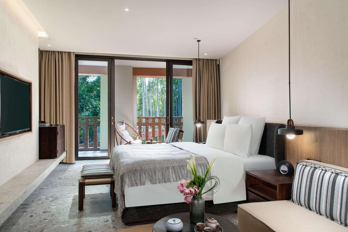 A hotel room with a bed, a sofa, a TV, a balcony, and a table with a vase and teapots.
