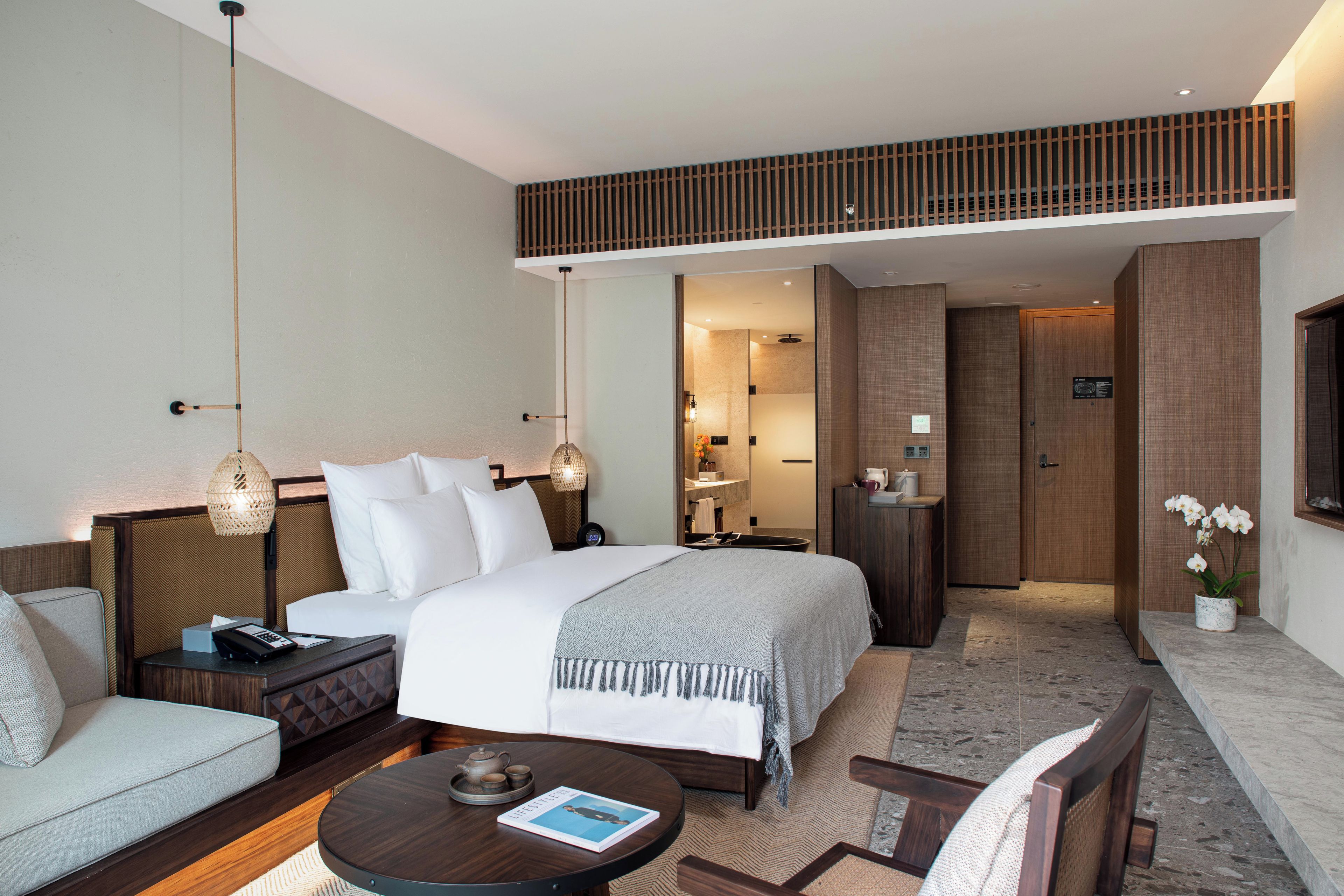 Habitación de hotel con cama, mesitas de noche, sofá, mesa de centro y baño visible a través de una puerta abierta.