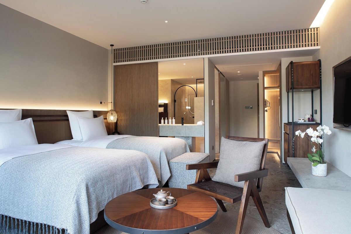 A hotel room with two beds, a sitting area with a round table, a TV, and a bathroom visible through an open door.